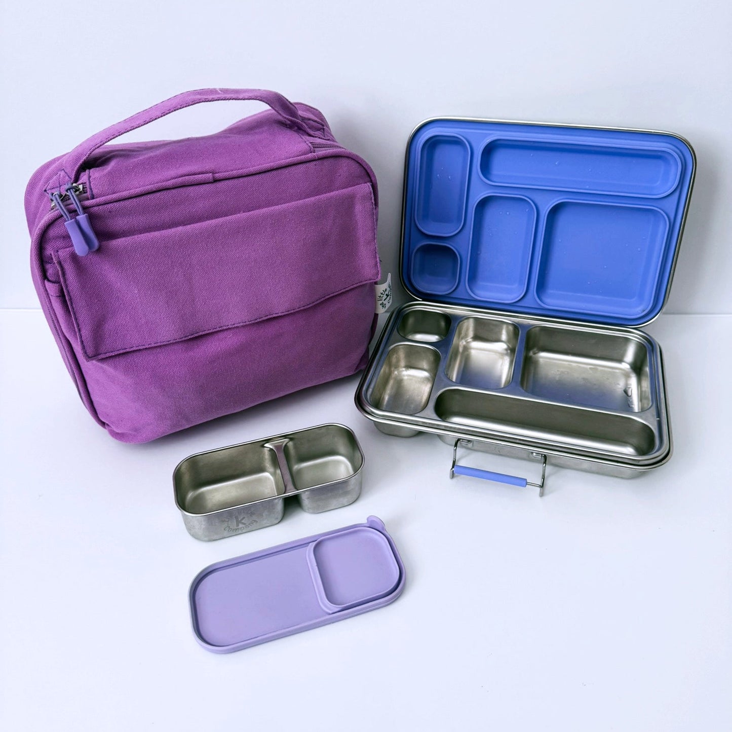 A purple fabric lunch bag shown next to an open stainless steel bento lunch box with a blue lid insert, plus two small compartment containers and a purple lid, all arranged on a clean white background.