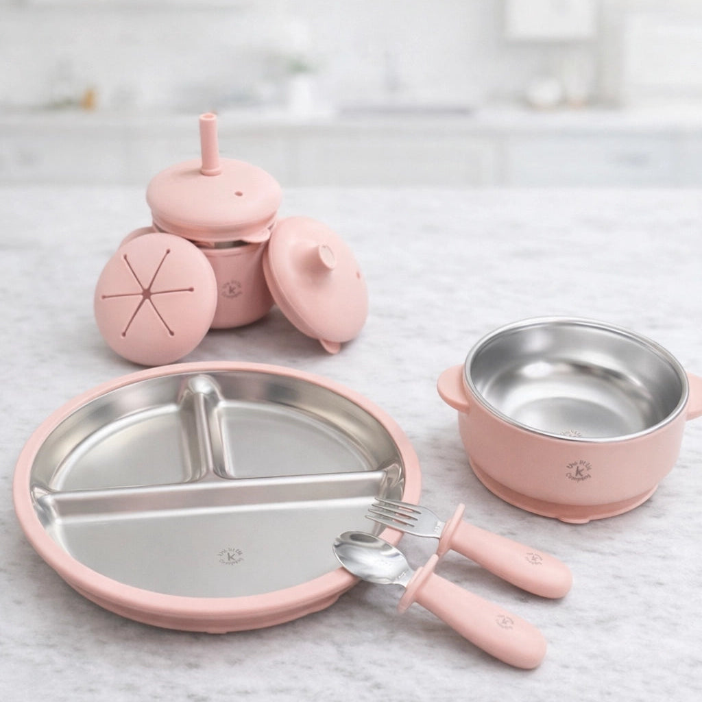Pink children's tableware set including a plate, bowl, utensils and convertible cup on a light gray surface.