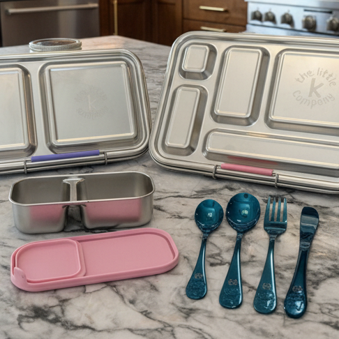 Set of stainless steel lunch containers and utensils on a marble countertop with a kitchen background.
