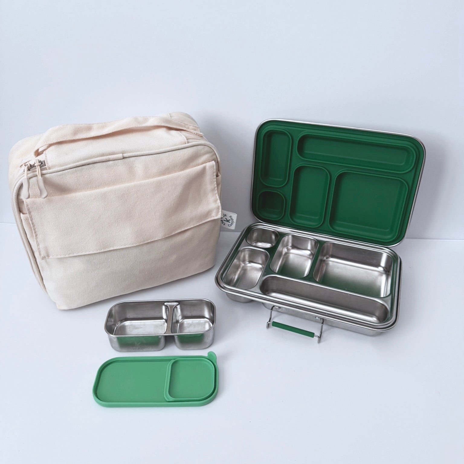 A stainless steel compartment lunch box set with a beige fabric bag, green silicone lids, and a green tray, arranged on a light gray background.
