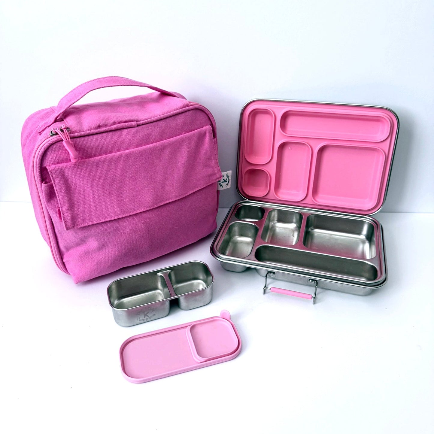 A pink lunch bag next to an open stainless steel bento box with pink compartments, plus two small matching pink and stainless steel lunch containers on a white background.