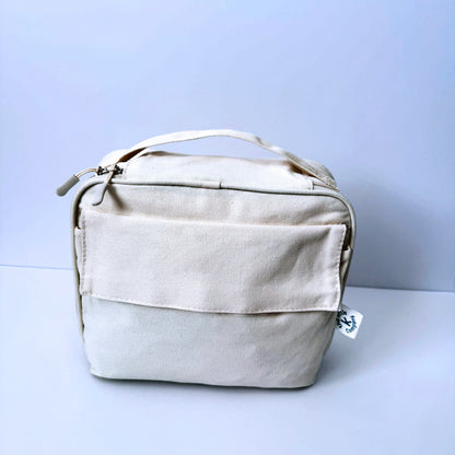A GOTS Certified Organic Cotton lunch bag in natural colour with a top handle, front pocket, and zipper closure, photographed against a light blue-gray studio background.